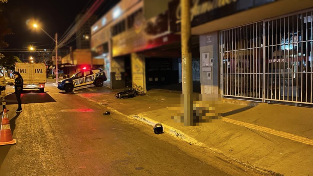 Imagem mostra um homem caído no chão ao lado de um poste. Mais a frente, está a motocicleta que ele dirigia antes de morrer. Foto ilustra a chamada: motociclista morre após colidir contra poste