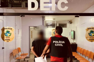 Foto mostra fachada da Delegacia e dois homens andando, de costas para a câmera. Um dos homens é um agente civil e outro o suspeito. foto ilustra chamada: Goianiense é suspeito de aplicar golpe em idosa do MG e causar prejuízo de R$ 30 mil