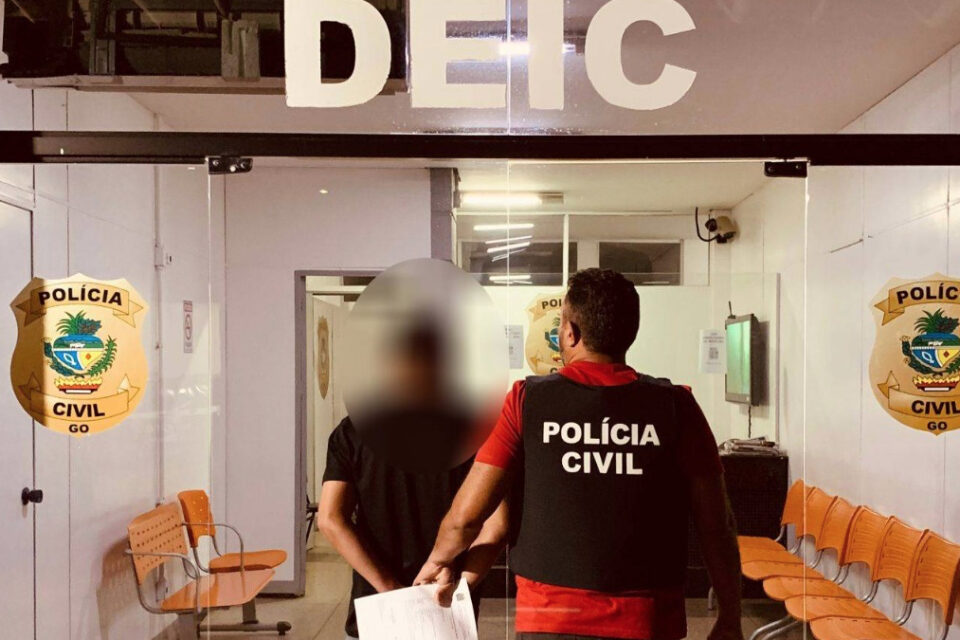 Foto mostra fachada da Delegacia e dois homens andando, de costas para a câmera. Um dos homens é um agente civil e outro o suspeito. foto ilustra chamada: Goianiense é suspeito de aplicar golpe em idosa do MG e causar prejuízo de R$ 30 mil
