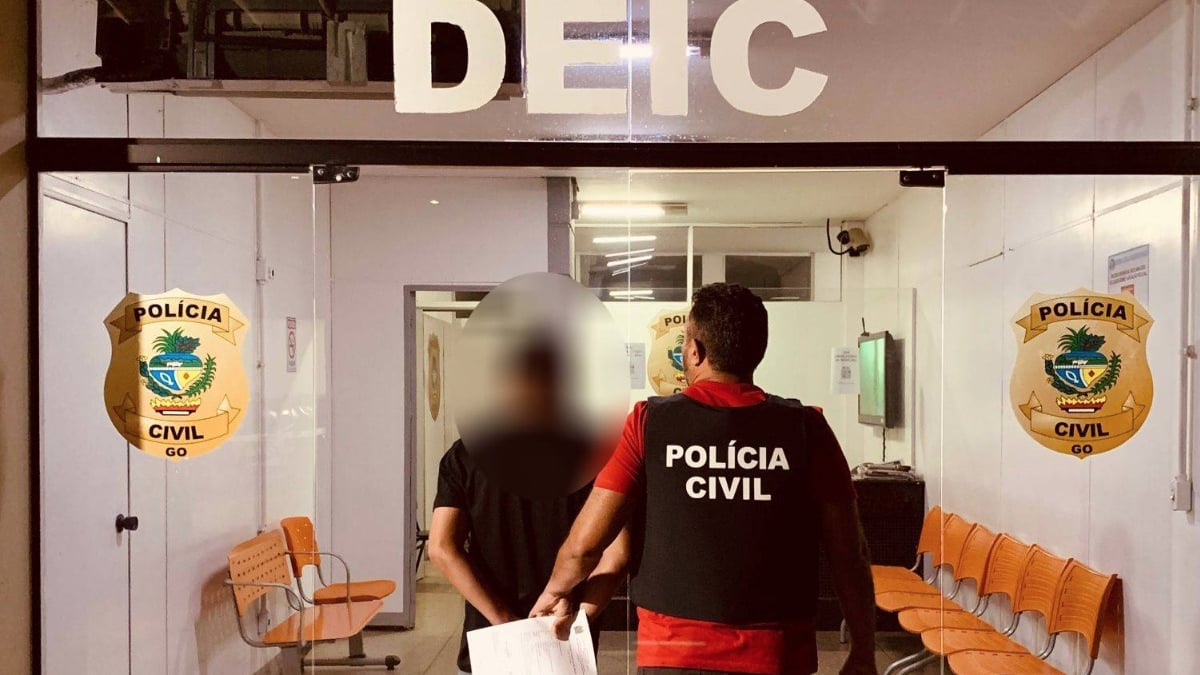 Foto mostra fachada da Delegacia e dois homens andando, de costas para a câmera. Um dos homens é um agente civil e outro o suspeito. foto ilustra chamada: Goianiense é suspeito de aplicar golpe em idosa do MG e causar prejuízo de R$ 30 mil