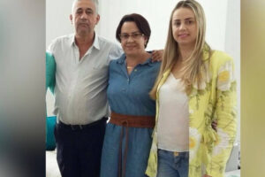 O pai à esquerda, a mãe no meio e a filha na outra extremidade posam para a foto. As duas mulheres sorriem e o homem se mantém sério.