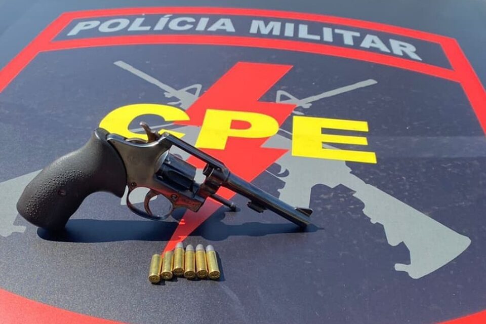 Revólver com munição em cima de um capô de viatura com logo do CPE da Polícia Militar