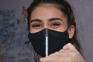 Seringa com vacina contra Covid-19 e em segundo plano mulher sorri