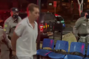 Foto mostra homem com camiseta branca, caminhando algemado, com militares atrás. Ao lado dele tem cadeiras azuis e ao fundo uma viatura da PM. Escondido em caverna, suspeito de espancar mulher por 4 horas é preso em Formosa