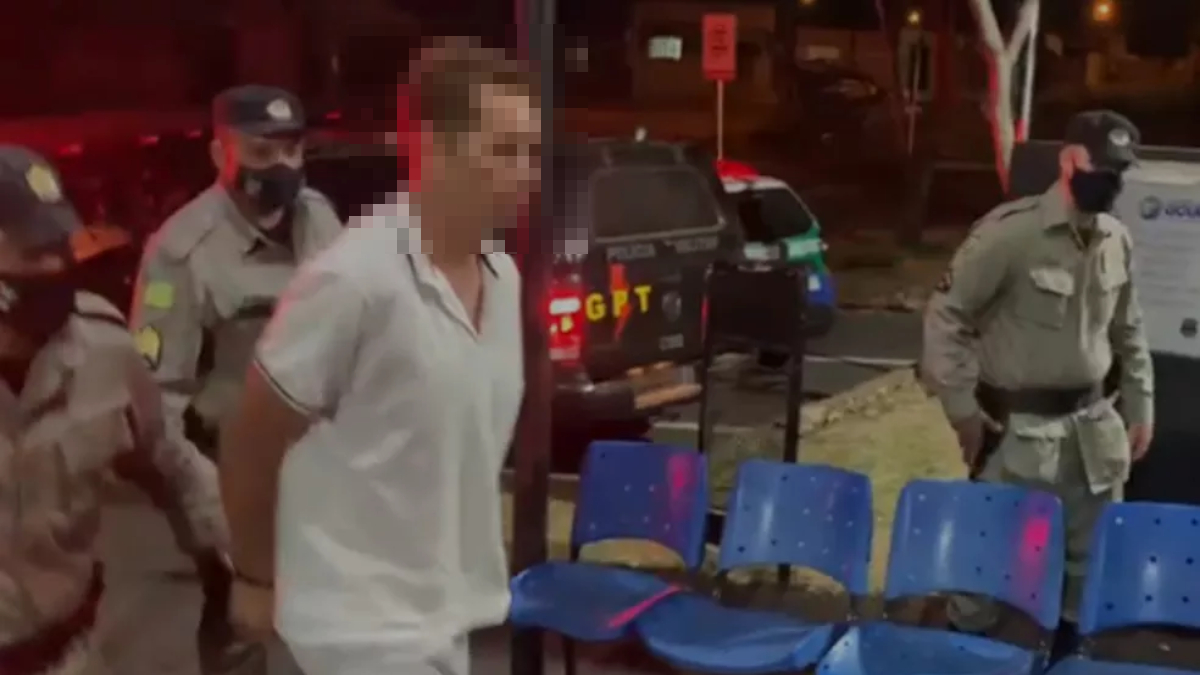 Foto mostra homem com camiseta branca, caminhando algemado, com militares atrás. Ao lado dele tem cadeiras azuis e ao fundo uma viatura da PM. Escondido em caverna, suspeito de espancar mulher por 4 horas é preso em Formosa