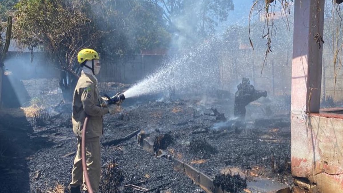 Bombeiro com mangueira de água controla chamas. Terreno está todo incendiado. Incêndio em vegetação de lote baldio atinge casa em Pirenópolis