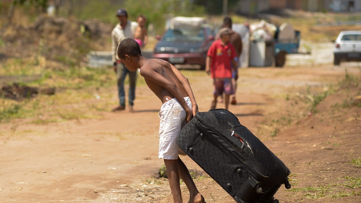 Criança puxa uma mala em terreno de chão batido a ocupação