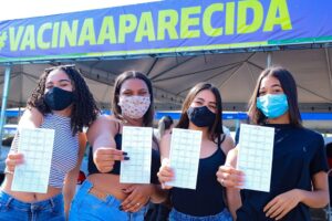 Quatro adolescentes de máscara mostram cartão de vacinação. Ao fundo faixa escrita: Vacina Aparecida