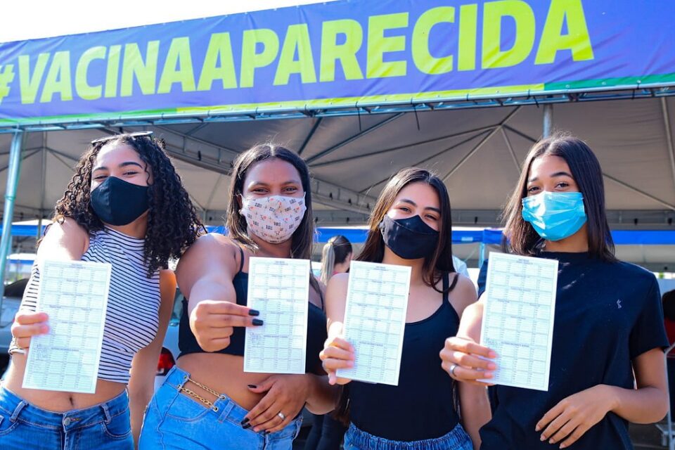 Quatro adolescentes de máscara mostram cartão de vacinação. Ao fundo faixa escrita: Vacina Aparecida