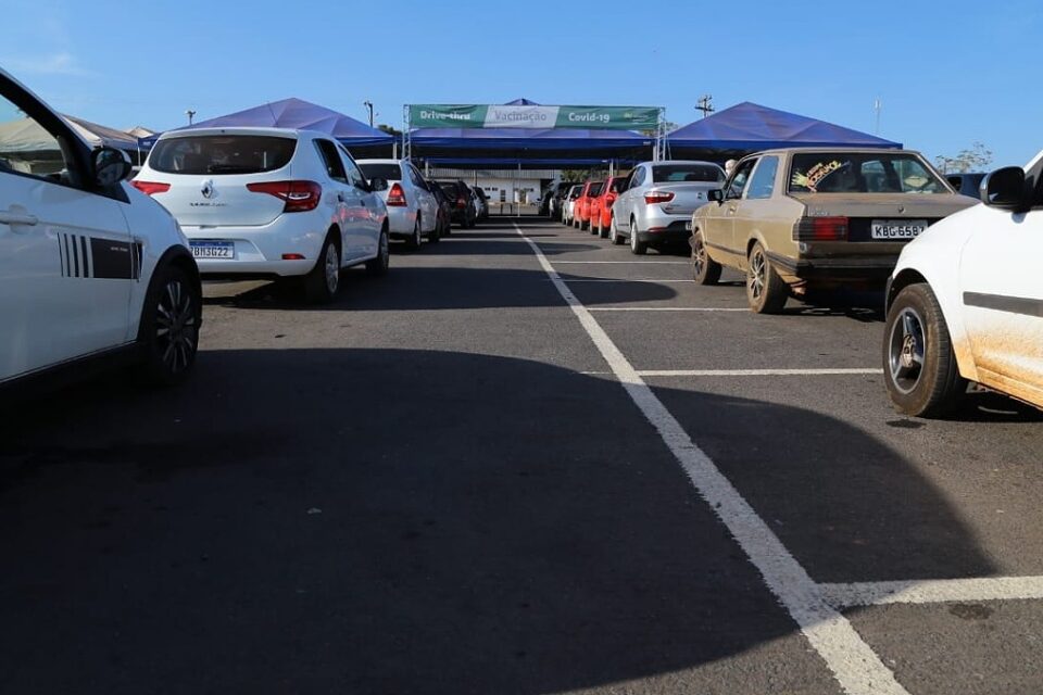 Fila de carros de dois lados em estacionamento. Ao fundo cartaz de vacinação em Aparecida de Goiânia