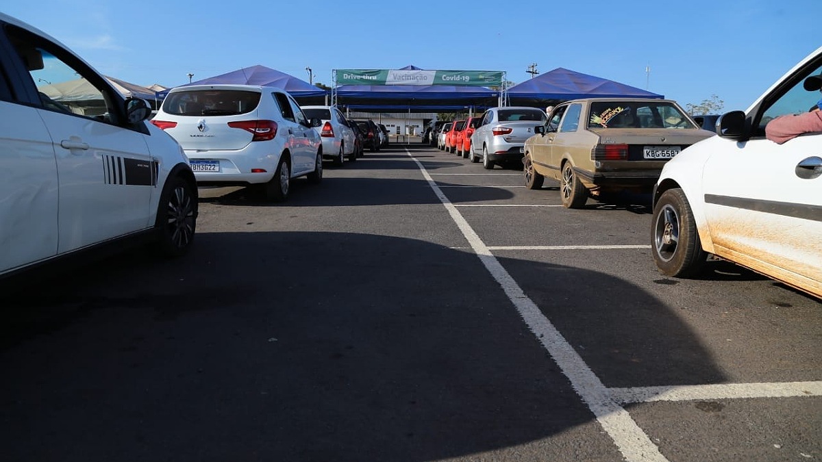 Fila de carros de dois lados em estacionamento. Ao fundo cartaz de vacinação em Aparecida de Goiânia