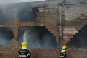 Dois bombeiros lançam água com mangueira em prédio que sai fumaça