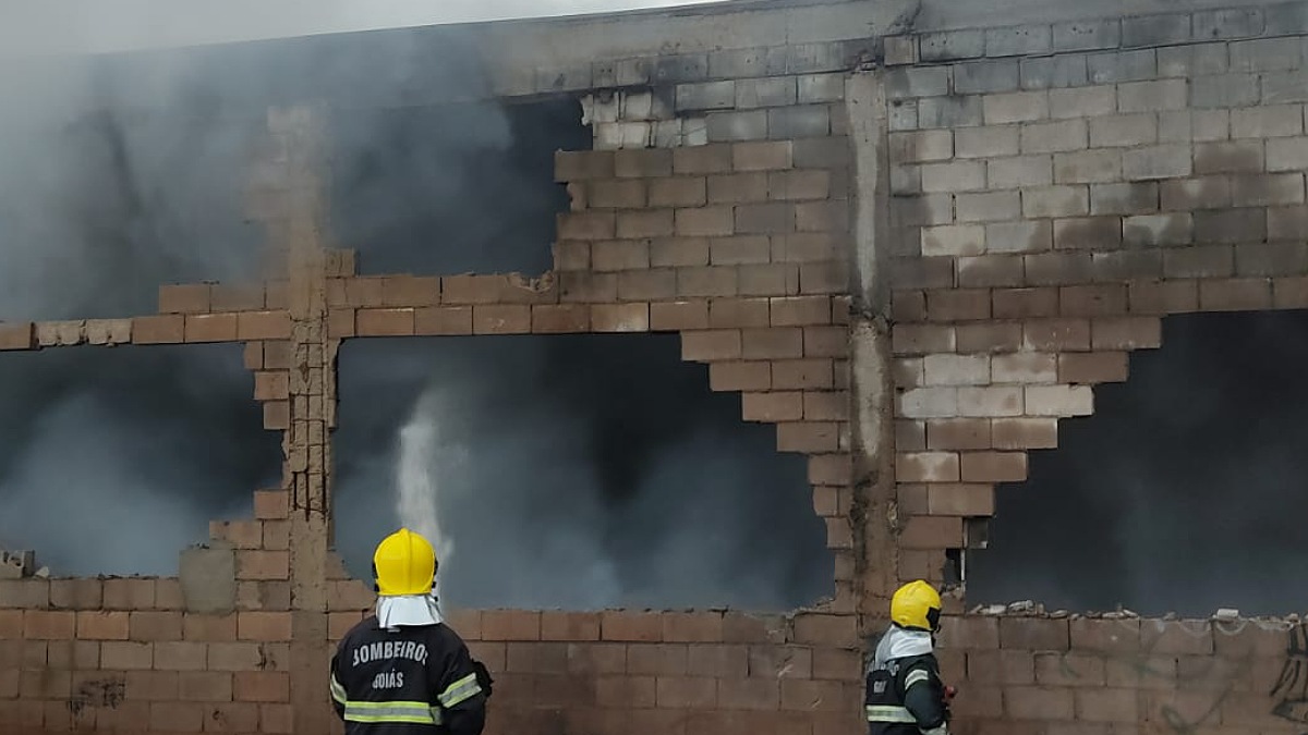 Dois bombeiros lançam água com mangueira em prédio que sai fumaça