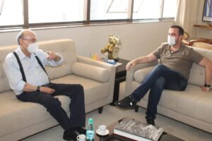 Henrique Meirelles e Gustavo Mendanha sentados frente a frente em sofás, com máscaras