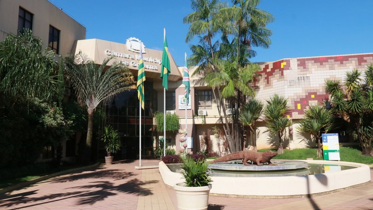 Entrada da Câmara Municipal de Goiânia com fonte e jacaré no primeiro plano e bandeiras no segundo