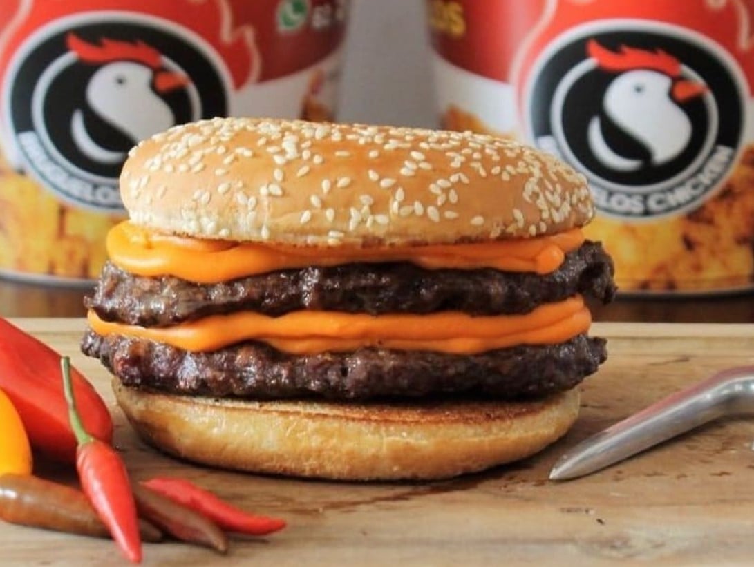 sanduíche com duas camadas de hambúrguer bovino e cheddar, com pimentas malaguetas no canto esquerdo. O sanduíche é do Bruguelos Chicken, opção de hambúrguer artesanal em Aparecida de Goiânia