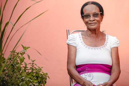 Imagem mostra a 1ª idosa vacinada contra Covid em Goiás. Ela usa óculos e está sentada em uma cadeira ao lado de uma planta.