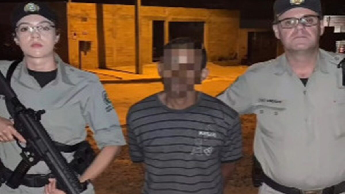 Suspeito de morder testículos do enteado aparece com as mãos para trás, sendo segurado por um policial militar.