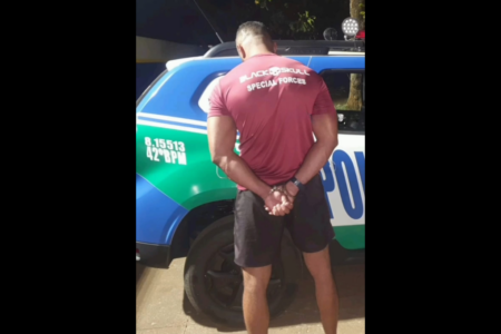 Um homem usando uma camiseta vermelha está de costas para a foto com a cabeça baixa. à sua frente tem uma viatura da Polícia Militar de Goiás.