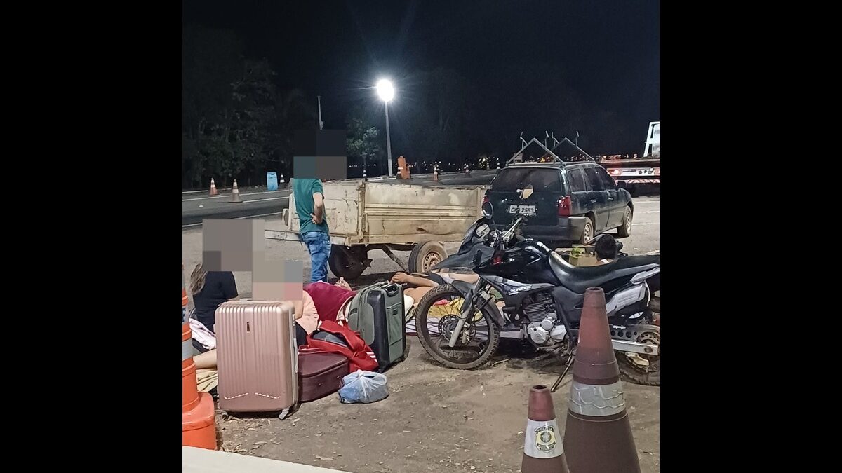 Foto colorida foi tirada à noite e mostra malas no chão, duas pessoas sentadas e uma em pé em meio à elas e uma moto preta parada ao lado