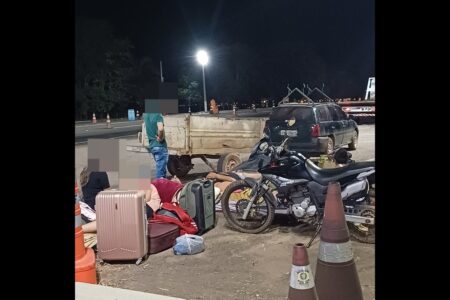 Foto colorida foi tirada à noite e mostra malas no chão, duas pessoas sentadas e uma em pé em meio à elas e uma moto preta parada ao lado