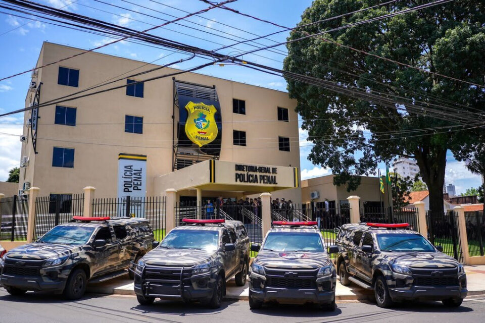 Imagem colorida mostra a entrada do prédio da Polícia Penal de Goiás com quatro viaturas pretas paradas na frente.