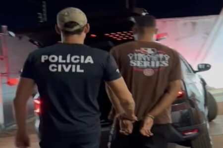 Imagem colorida mostra um policial civil do lado esquerdo. Ele está de costas, usando uma camiseta preta e um boné verde escuro. Do lado direito, um homem de camiseta verde, algemado, sendo levado pelo agente.