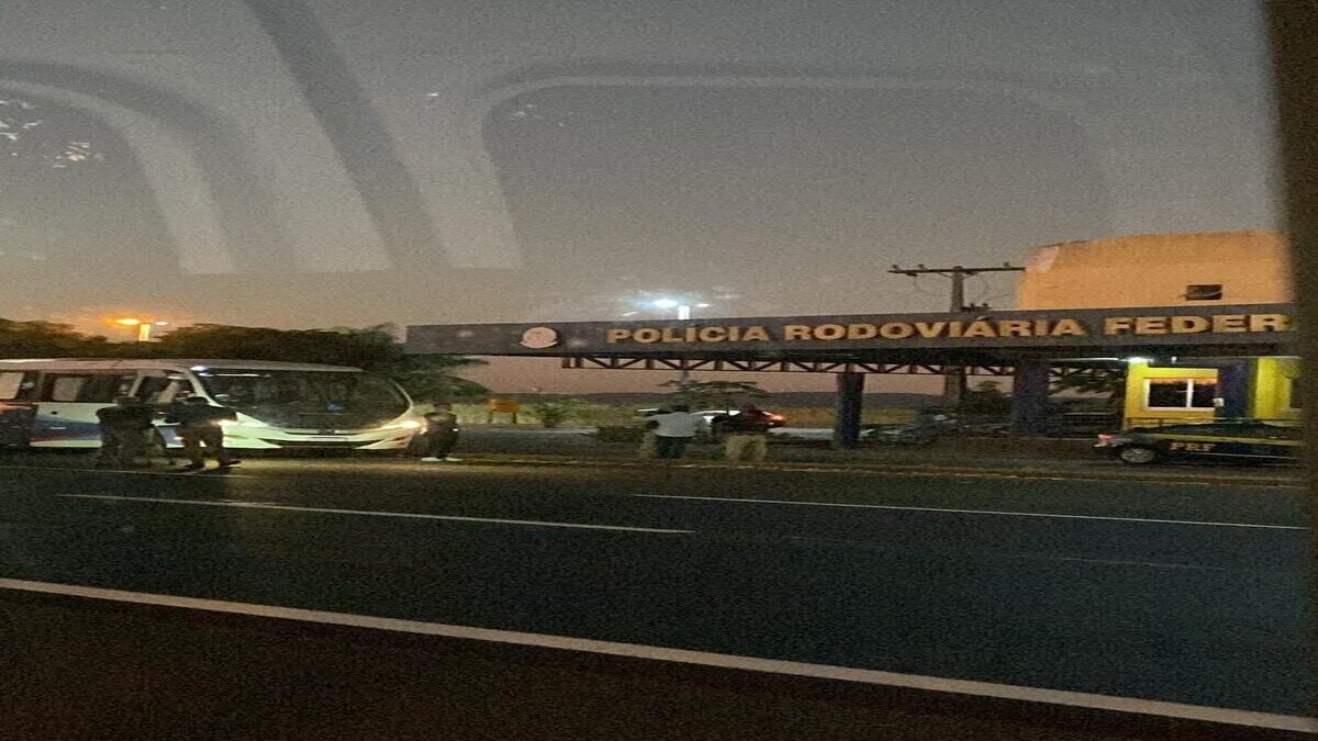Imagem colorida mostra um ônibus parado em frente a um posto da Polícia Rodoviária Federal.