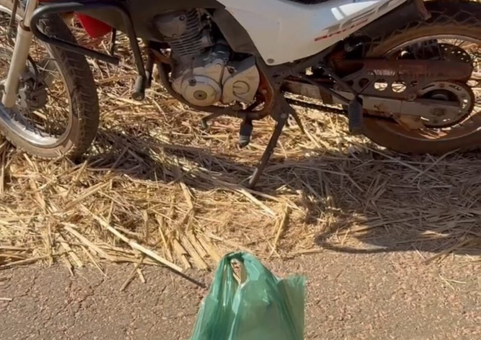 Imagem colorida mostra uma moto vermelha, preta e branca parada. à sua frente uma sacola verde.