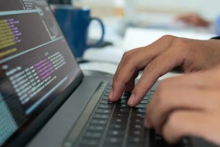 Imagem colorida mostra parte de um notebook preto e duas mãos como se estivessem digitando.