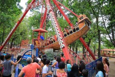 Imagem colorida mostra diversas pessoas no Parque de Diversão em frente a um brinquedo chamado barco Viking.