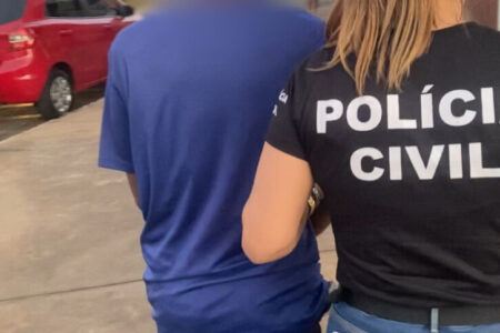 Imagem colorida mostra um homem de camiseta azul sendo levado por uma agente da Polícia Civil.