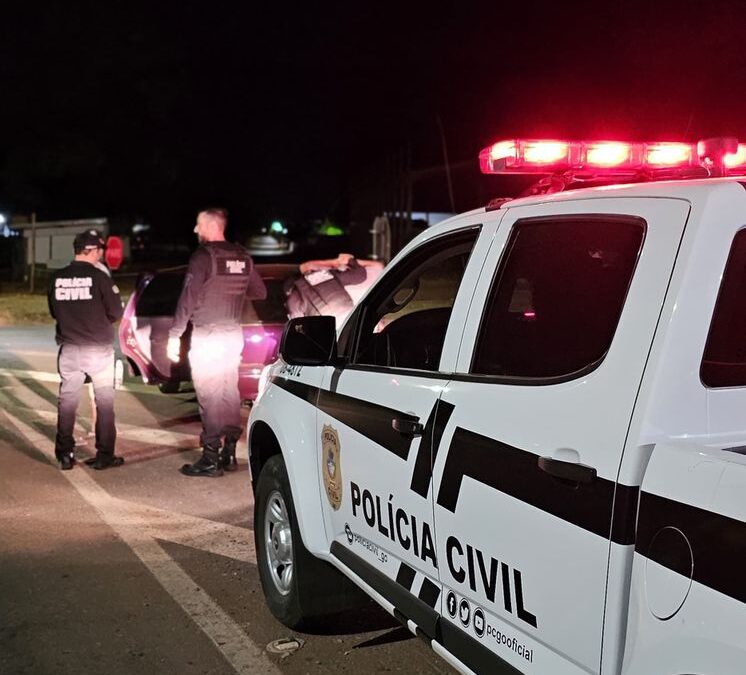 Imagem mostra uma viatura da Polícia Civil em primeiro plano e agentes ao fundo.