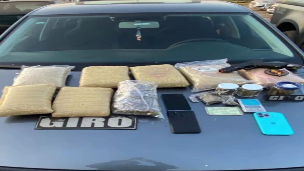 Imagem colorida mostra o capô de uma viatura da Polícia Militar. Em cima dela, quatro pacotes de drogas na cor marrom, um celular e notas de dinheiro.