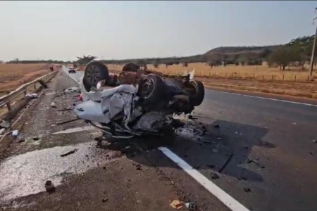 Imagem colorida mostra um carro branco destruído após um acidente em uma rodovia.