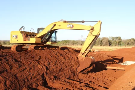 Imagem colorida mostra uma máquina retroescavadeira em um terreno de terra