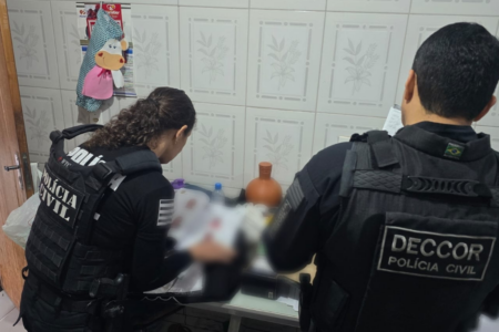 Imagem colorida mostra dois agentes da Polícia Civil em frente a uma mesa verificando e lendo documentos.