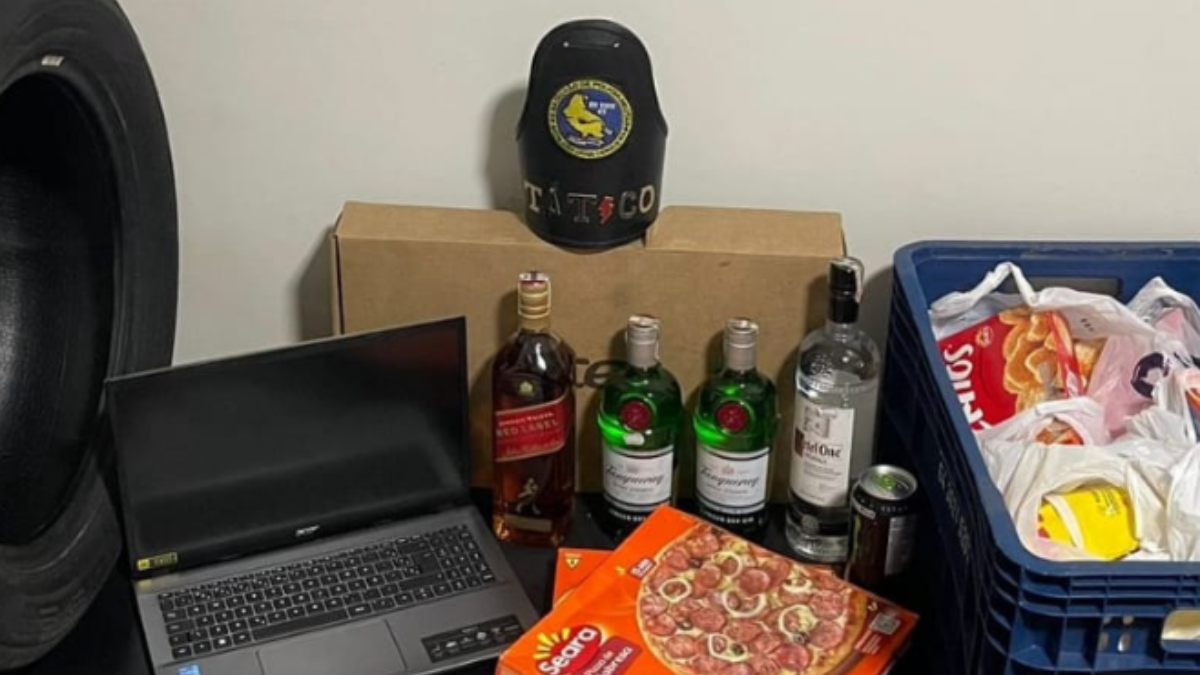 Imagem colorida mostra itens apreendidos na casa de um suspeito. Um notebook, uma garrafa de whisky, duas de gin e uma embalagem de pizza congelada estão entre os itens
