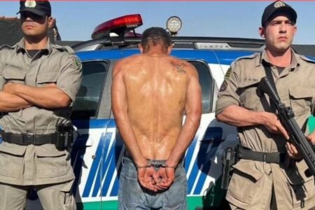 Foto colorida mostra homem preso diante de viatura e policiais militares