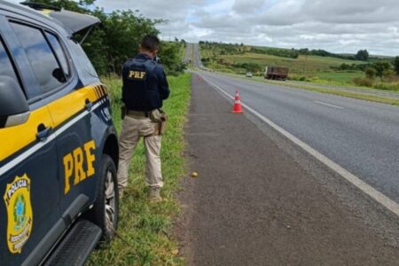 Imagem colorida mostra um agente da PRF ao lado de uma viatura parados em uma rodovia.