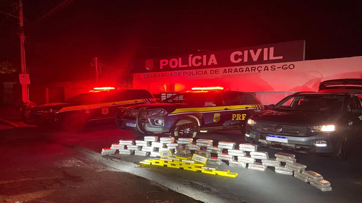 Imagem colorida mostra diversos tabletes de drogas em frente à viaturas da Polícia Rodoviária Federal.