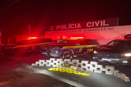 Imagem colorida mostra diversos tabletes de drogas em frente à viaturas da Polícia Rodoviária Federal.