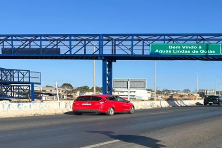 Imagem colorida mostra um carro vermelho passando por uma rodovia embaixo de uma passarela.