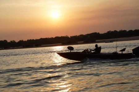 Imagem colorida mostra um barco em um rio com o pôr do sol ao fundo.