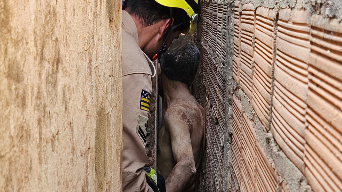 Imagem colorida mostra um homem sem camisa entre duas paredes de tijolo e um bombeiro realizando o resgate.