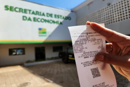 Imagem colorida mostra a fachada da Secretaria de Economia e uma mão segurando uma nota fiscal.