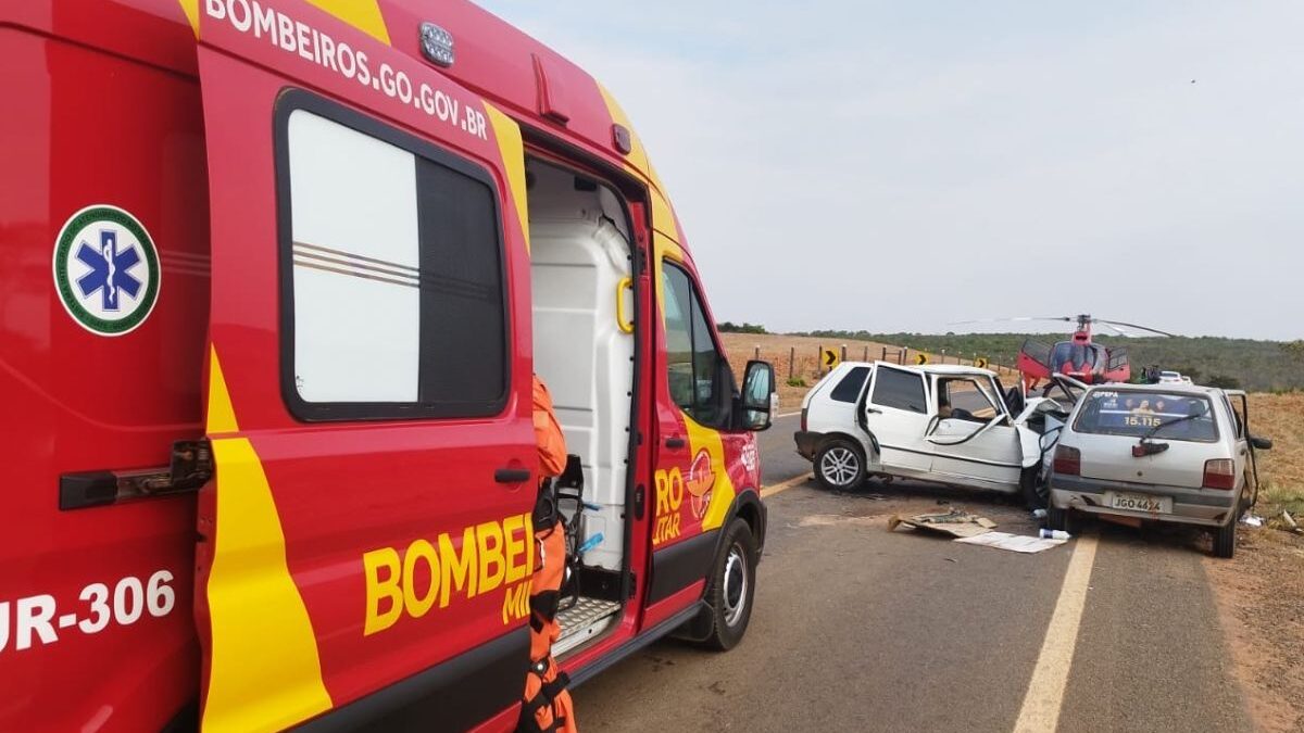 Imagem colorida mostra em primeiro plano uma viatura do Corpo de Bombeiros com a porta aberta e, mais ao fundo, um carro branco com a lateral amassada após um acidente.