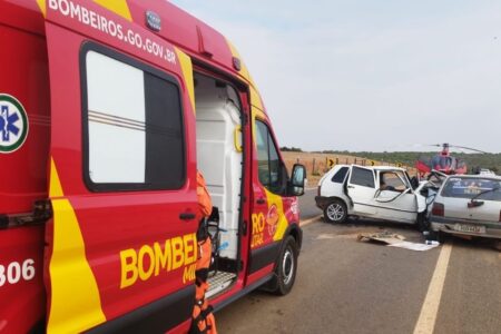 Imagem colorida mostra em primeiro plano uma viatura do Corpo de Bombeiros com a porta aberta e, mais ao fundo, um carro branco com a lateral amassada após um acidente.