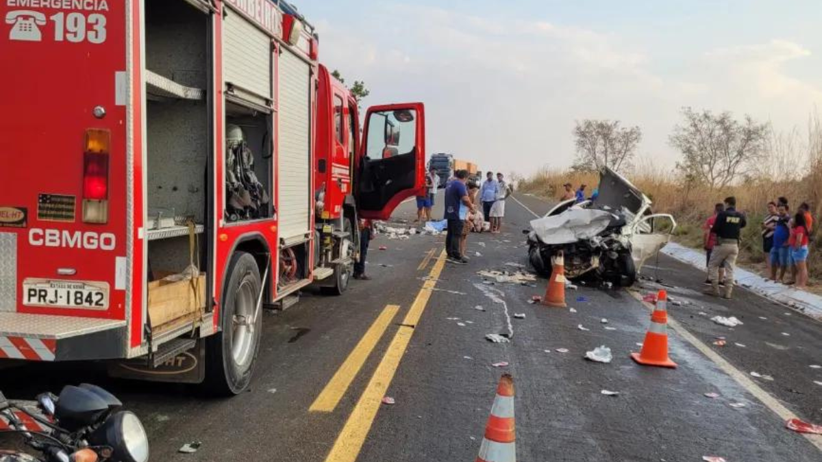 Imagem colorida mostra, em primeiro plano, um caminhão do Corpo de Bombeiros e, ao fundo, um veículo branco destruído após um grave acidente,