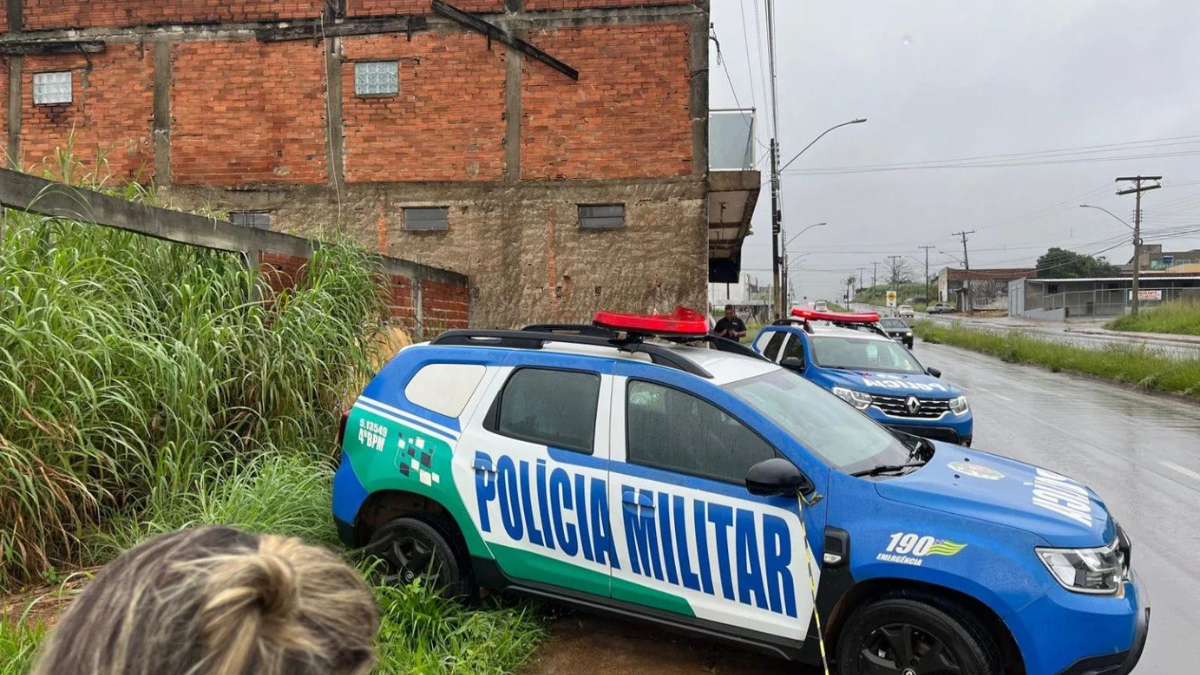 Imagem colorida mostra uma viatura da Polícia Militar parada em uma área com mato alto, em frente a uma casa com parede de tijolos.